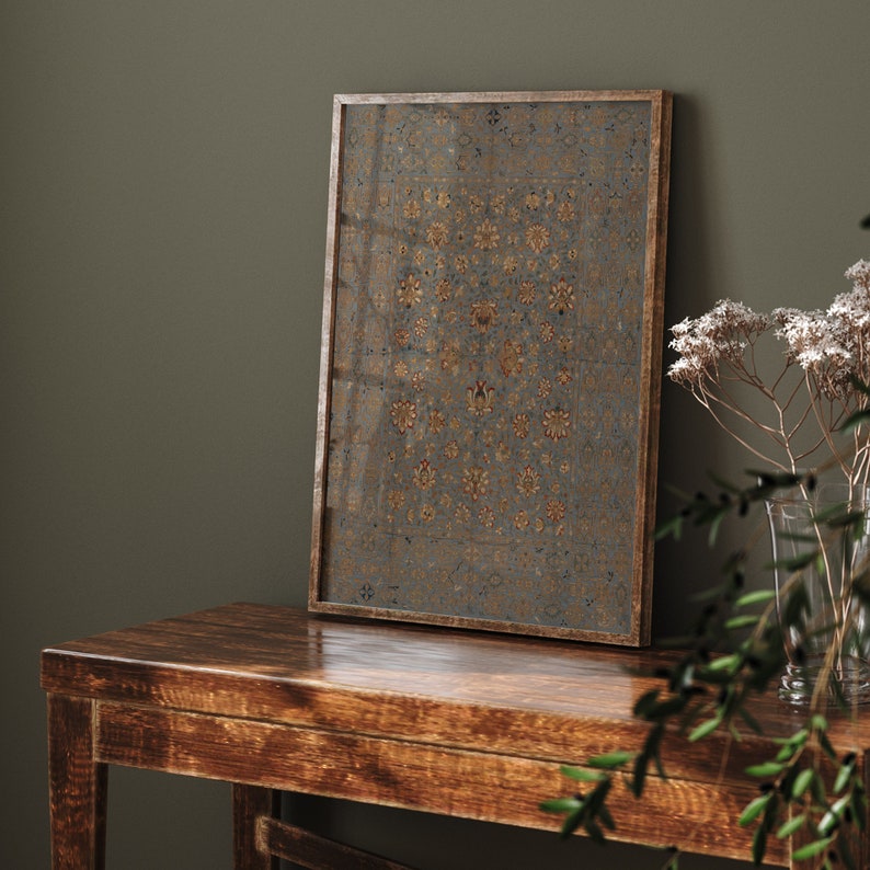 May include: A framed print of a vintage rug with a geometric pattern in shades of gold, blue, and red. The print is hanging on a dark green wall above a wooden table.