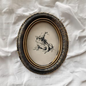 May include: An oval framed art print with a pen and ink drawing of a frog in a witch's hat. The frame has a dark, textured outer edge and a gold inner rim. The text "ATLAS VINTAGE" is at the bottom of the print.