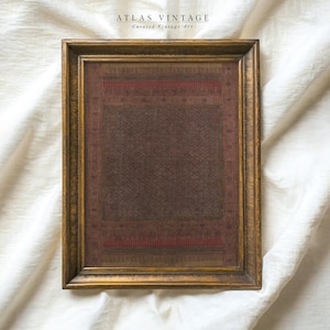 May include: A framed vintage textile with a detailed geometric pattern in shades of brown and red. The frame is a warm gold color. The text "ATLAS VINTAGE Curated Vintage Art" is at the top.