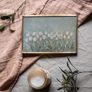 May include: A gold-framed painting of white tulips on a light blue background. The painting sits on a pink cloth, with a lit candle in a glass jar and sprigs of greenery. The painting's style is reminiscent of impressionism.