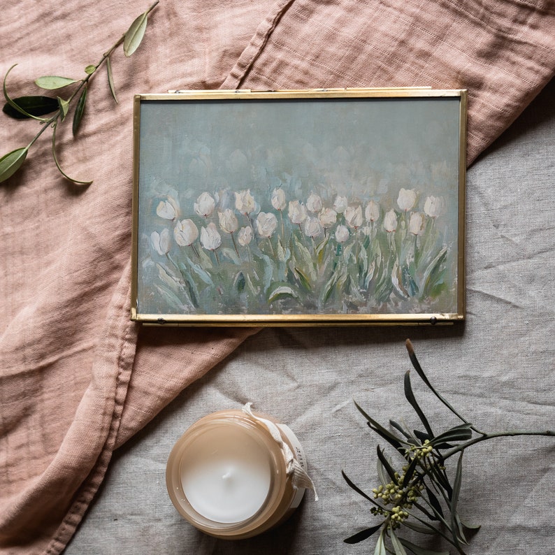 May include: A gold-framed painting of white tulips on a light blue background. The painting sits on a pink cloth, with a lit candle in a glass jar and sprigs of greenery. The painting's style is reminiscent of impressionism.