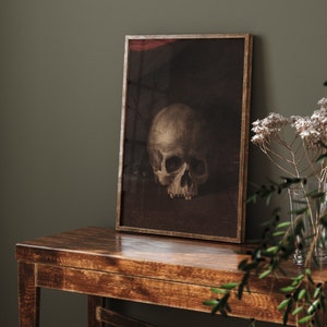 May include: A framed print of a still life featuring a human skull in a dark, muted color palette. The skull is positioned on a dark surface, with a subtle red background.