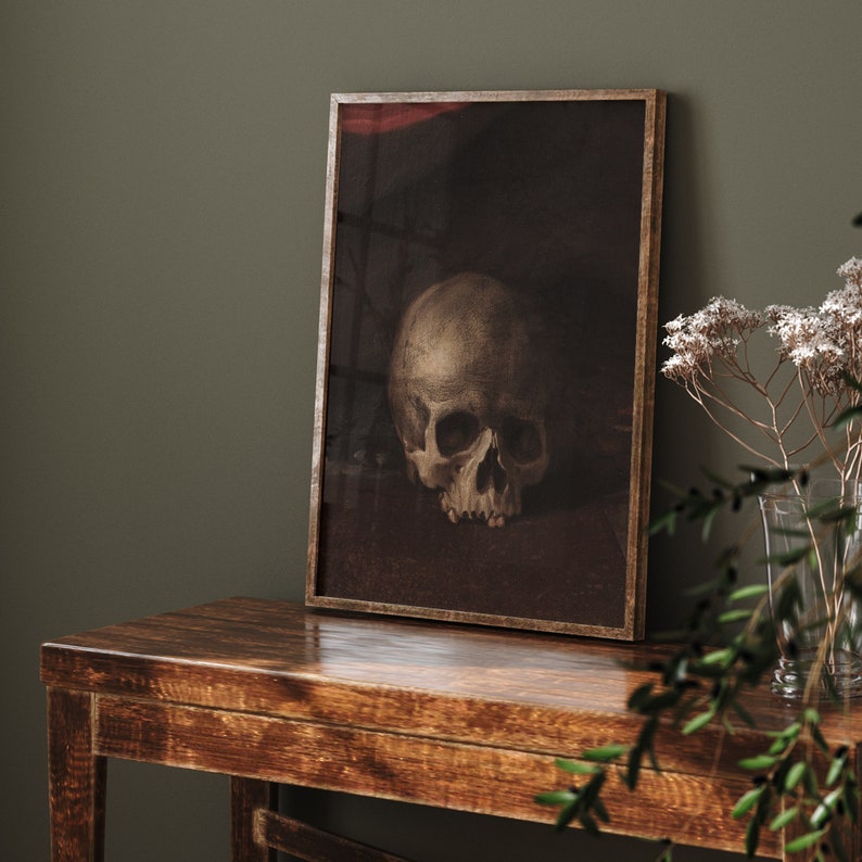 May include: A framed print of a still life featuring a human skull in a dark, muted color palette. The skull is positioned on a dark surface, with a subtle red background.