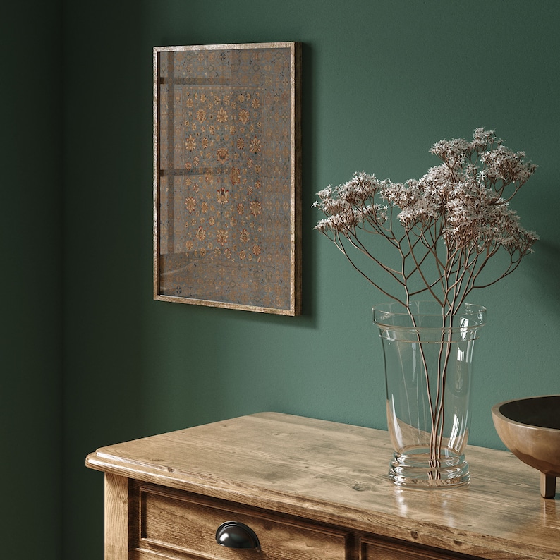 May include: A wooden dresser with three drawers, a vase with dried flowers, and a framed artwork with a geometric pattern. The dresser is made of light brown wood and has a natural finish. The vase is clear glass and the flowers are white. The artwork is framed in gold and has a dark background with a gold pattern.