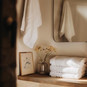 May include: A bathroom scene featuring a stack of three folded white towels, a framed lighthouse illustration, and a clear glass vase with dried flowers. A white towel hangs on the wall, and a mirror is visible.