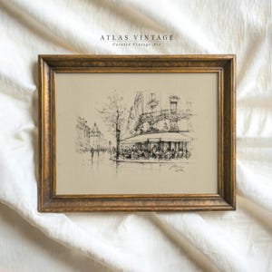 May include: A framed vintage sketch of a European cafe scene. The artwork, in black and white, depicts a street with buildings, a tree, and people seated at tables under an awning. The frame is gold-toned, and the text "ATLAS VINTAGE Curated Vintage Art" is visible.