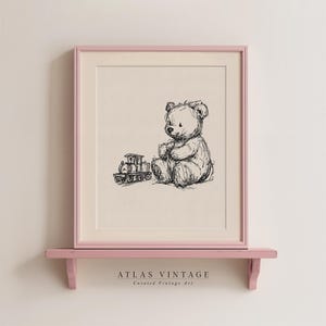 May include: A framed monochrome sketch of a teddy bear and a toy train. The artwork is displayed on a pink shelf with the text "ATLAS VINTAGE Curated Vintage Art" below the image. The frame is a pale pink colour.