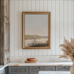 May include: A gold framed painting of a landscape with a sailboat in the distance. The painting is hanging on a white paneled wall above a white countertop.