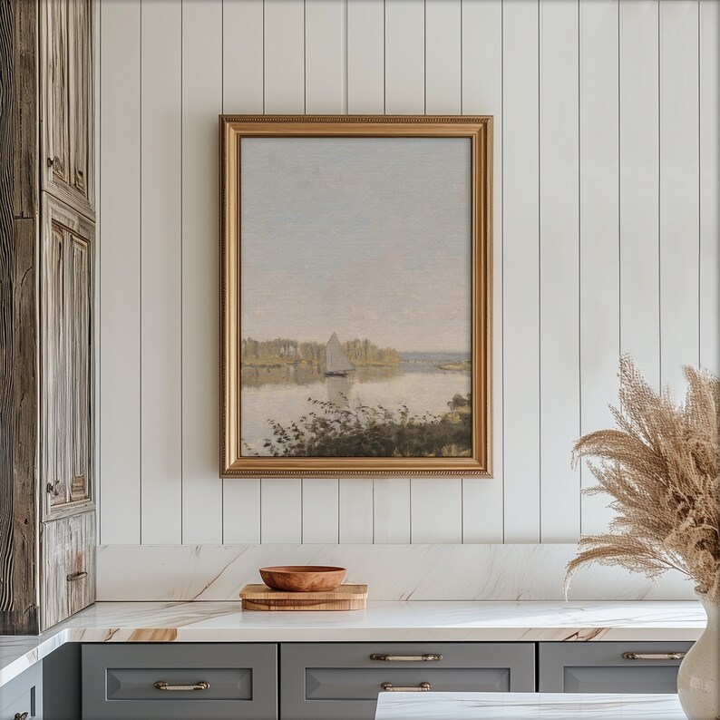 May include: A gold framed painting of a landscape with a sailboat in the distance. The painting is hanging on a white paneled wall above a white countertop.