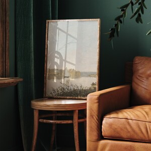 May include: A framed landscape painting of a lake with trees and a sailboat in the distance. The painting is hanging on a wall in a room with a brown leather armchair and a wooden side table.