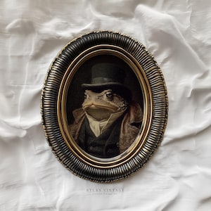 Puede incluir: Retrato ovalado enmarcado de una rana vestida con sombrero de copa negro, traje y abrigo. El marco es negro con detalles dorados. La obra de arte está etiquetada "ATLAS VINTAGE" en la parte inferior.