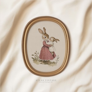 May include: A vintage oval framed print of two brown rabbits, one holding the other. The larger rabbit is wearing a pink dress with white polka dots. The smaller rabbit is wearing a white dress. The print is on a cream background and the frame is gold.