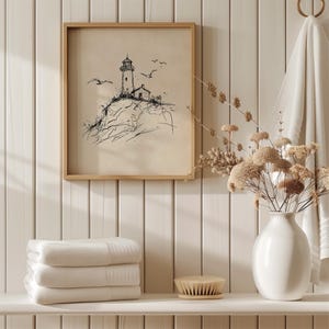 May include: A framed black and white lighthouse sketch with seagulls, set against a neutral background. The artwork is displayed above a shelf holding folded white towels, a brush, and a white vase with dried flowers. The scene evokes a coastal theme.