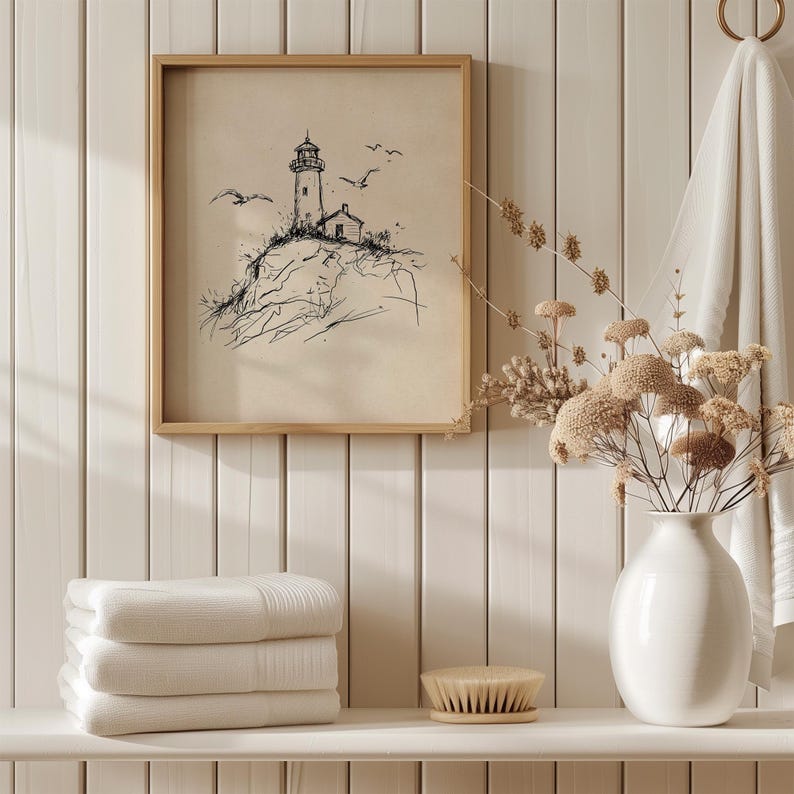 May include: A framed black and white lighthouse sketch with seagulls, set against a neutral background. The artwork is displayed above a shelf holding folded white towels, a brush, and a white vase with dried flowers. The scene evokes a coastal theme.