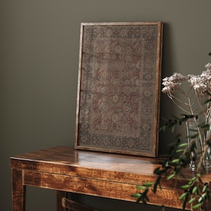 May include: A framed print of a vintage rug with intricate patterns in shades of red, brown, and beige. The frame is a dark brown wood and the print is sitting on a wooden table.
