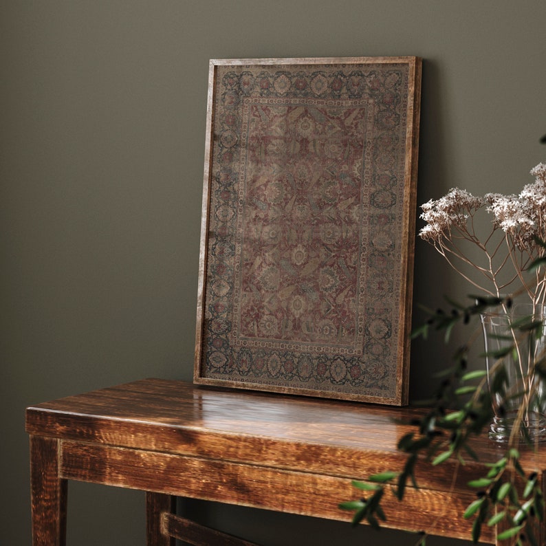 May include: A framed print of a vintage rug with intricate patterns in shades of red, brown, and beige. The frame is a dark brown wood and the print is sitting on a wooden table.