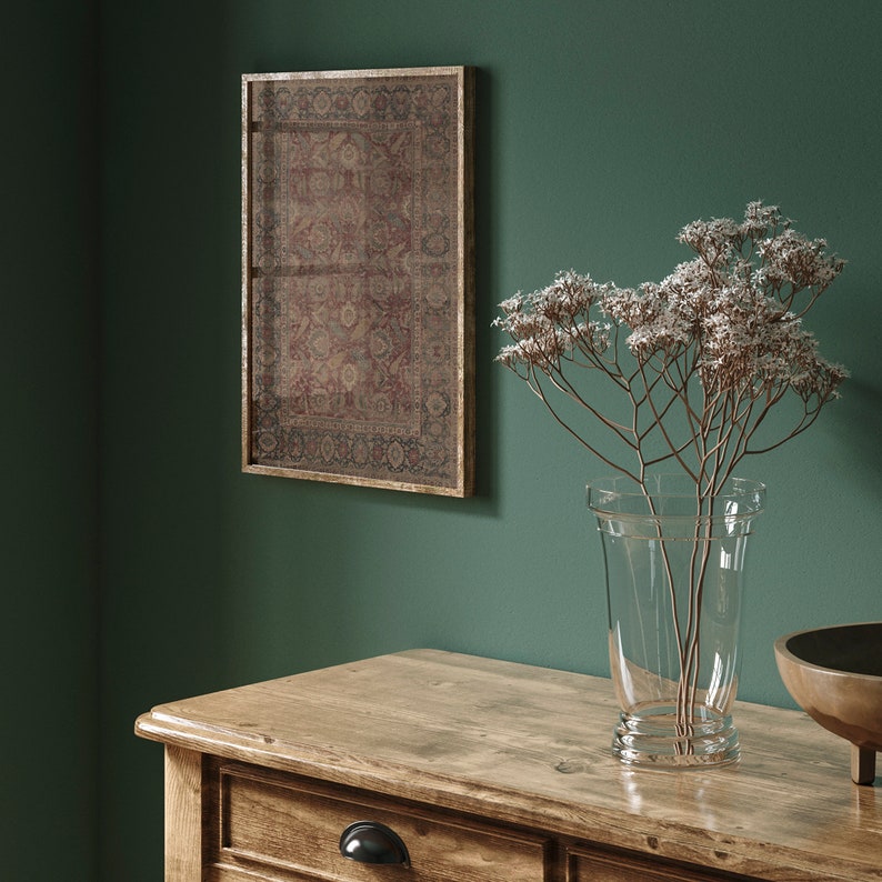 May include: A wooden table with a drawer and a vase of dried flowers. The vase is clear glass and the flowers are white. There is a framed picture of a rug hanging on the wall behind the table.