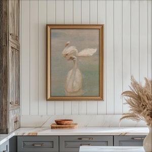May include: A framed painting of two white swans swimming in a lake. The painting has a gold frame and a light blue background.