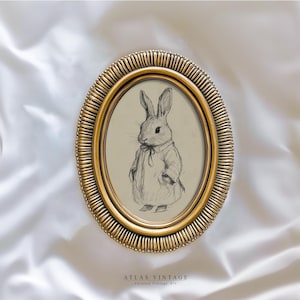 May include: A black and white drawing of a rabbit wearing a dress is framed in a gold oval frame.