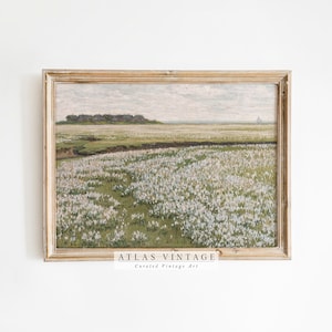 Puede incluir: Una pintura vintage enmarcada que representa un paisaje con un campo de flores blancas en primer plano, un pequeño pueblo en la distancia y un cielo nublado. El marco es de madera clara. La pintura se titula "ATLAS VINTAGE Arte Vintage Curado".