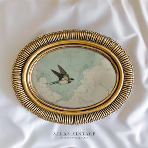 Puede incluir: Una obra de arte enmarcada ovalada de color dorado que representa un pájaro en vuelo contra un cielo nublado. El pájaro tiene marcas negras, blancas y azules. El marco tiene un diseño texturizado y acanalado. El texto "ATLAS VINTAGE" y "Curated Vintage Art" están en la parte inferior.