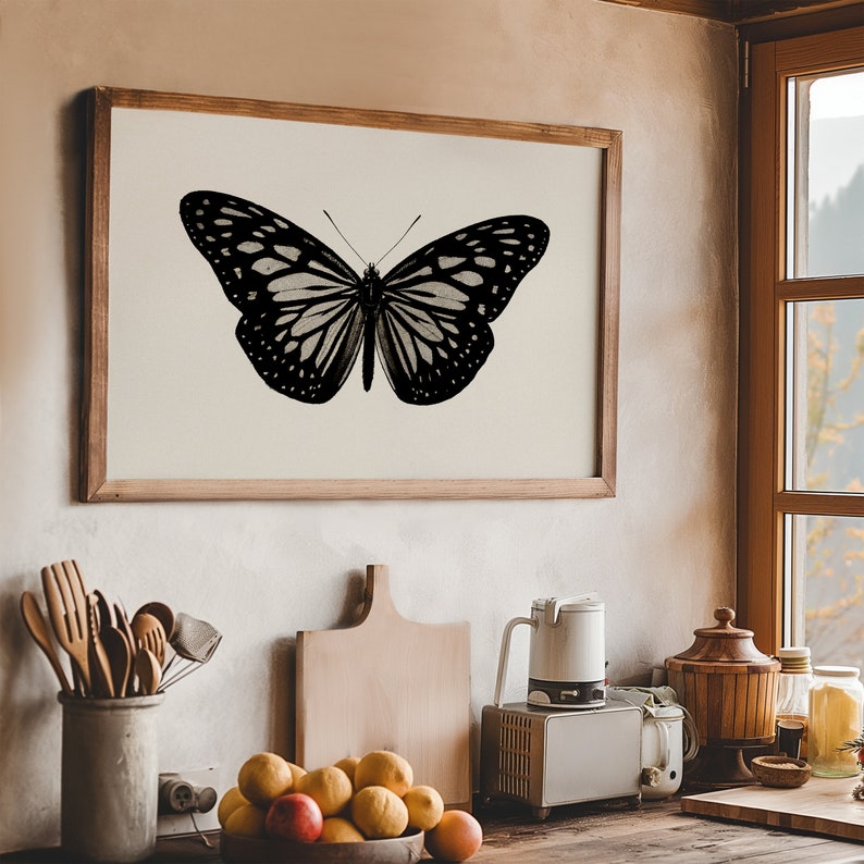 K&ouml;nnte beinhalten: Eine Schwarz-Wei&szlig;-Illustration eines Schmetterlings in einem Holzrahmen. Der Schmetterling ist auf einem cremefarbenen Hintergrund zentriert. Der Rahmen h&auml;ngt an einer Wand in einer K&uuml;chenumgebung.