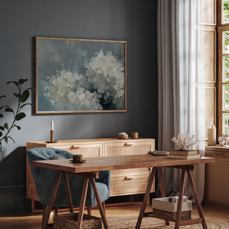 May include: A blue velvet armchair with a wooden frame sits in front of a wooden dresser with three drawers. A wooden table with a trestle base sits in front of the chair. A framed painting of white flowers on a blue background hangs on the wall behind the dresser.