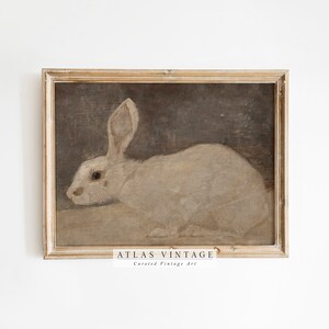 Puede incluir: Una pintura vintage enmarcada de un conejo blanco con ojos marrones, sobre un fondo marr&oacute;n oscuro. El marco es de madera clara con un acabado blanqueado. La pintura est&aacute; titulada "ATLAS VINTAGE Arte vintage seleccionado".