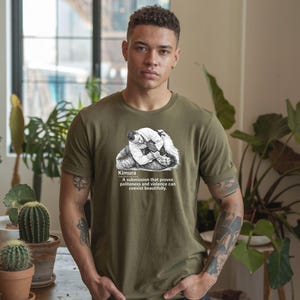 May include: Olive green t-shirt featuring a black and white illustration of a grappling hold, with the word "Kimura" and a description below. The text reads: "A submission that proves politeness and violence can coexist beautifully."