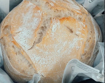 May include: A freshly baked loaf of sourdough bread, still warm from the oven, is wrapped in white parchment paper. The bread has a golden brown crust and a soft, airy interior.