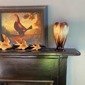 May include: A brown and white ceramic vase with a unique swirl design sits on a black mantelpiece.  Three small bird figurines are on the mantelpiece in front of a framed picture of a rooster.