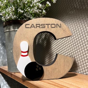Pu&ograve; includere: Lettera C in legno con un birilli e una palla da bowling, dipinti in bianco e nero con strisce rosse. La lettera &egrave; dipinta in un motivo a venature di legno marrone chiaro. La lettera ha il nome "CARSTON" inciso.