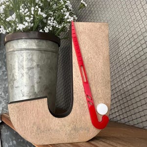 May include: Wooden letter J with a red hockey stick and white puck. The letter is painted in a light brown color with a distressed finish.