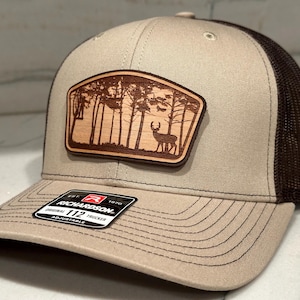 May include: A beige and brown trucker hat with a wood patch featuring a silhouette of a deer in a forest. The patch is attached to the front of the hat. The hat has a mesh back and an adjustable strap. The hat has the text "Richardson 112 Trucker Adjustable" on the front.