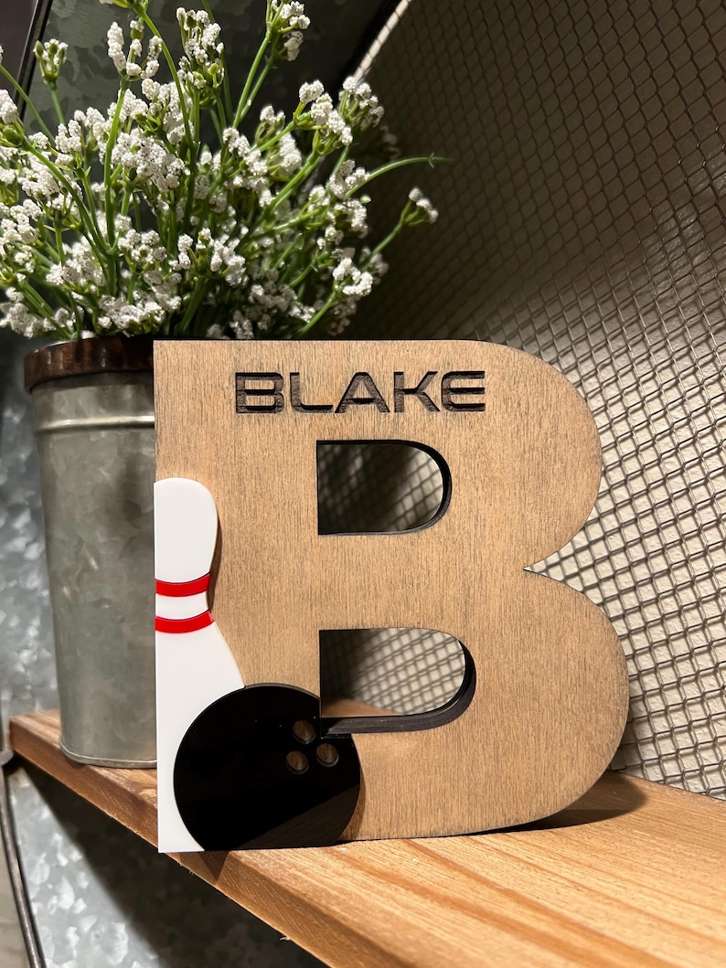 Pu&ograve; includere: Lettera B in legno con il nome "BLAKE" inciso su di essa. La lettera &egrave; decorata con un birilli da bowling bianco e una palla da bowling nera.