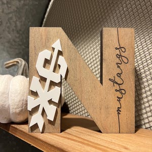 May include: Wooden letter N with a white snowflake and arrow design on the left side. The right side of the letter has the word "Mustangs" written in cursive.