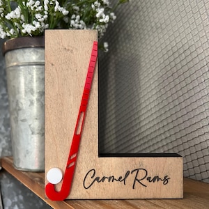 May include: Wooden letter L with a red field hockey stick and white ball, and the text "Carmel Rams" engraved on the bottom.