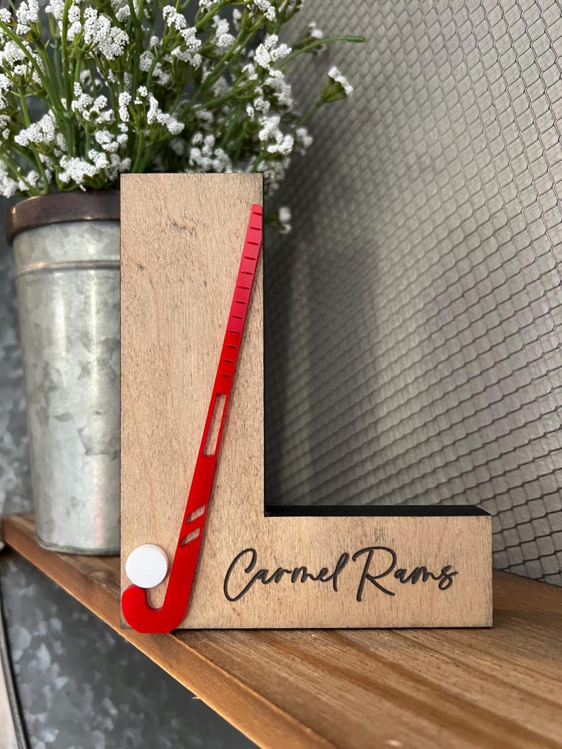 May include: Wooden letter L with a red field hockey stick and white ball, and the text "Carmel Rams" engraved on the bottom.