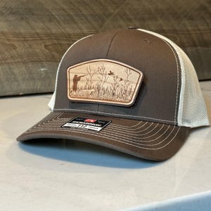 May include: A brown and white trucker hat with a wooden patch featuring a hunting scene. The patch shows a hunter, birds, and tall grass. The hat has a curved brim and a mesh back.