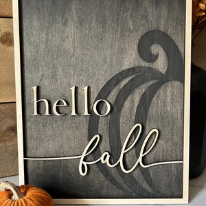 Może przedstawiać: Drewniany znak z ciemnoszarym tłem i jasną drewnianą ramą. Znak głosi "hello fall" kursywą. Stylizowana dynia jest wycięta z drewna za tekstem.