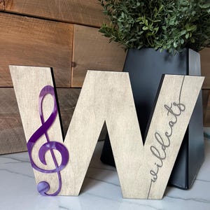 Könnte beinhalten: Holzbuchstabe "W" mit einem lila Violinschlüssel und dem Wort "Wildcats" in Schreibschrift. Der Buchstabe ist hellbraun mit dunklem Rand. Der Violinschlüssel hat eine leuchtende lila Farbe. Der Hintergrund umfasst eine schwarze Vase und Grün.