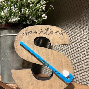 May include: Wooden letter S with the word "Spartans" carved into it. A blue hockey stick with a white puck is placed on the letter.