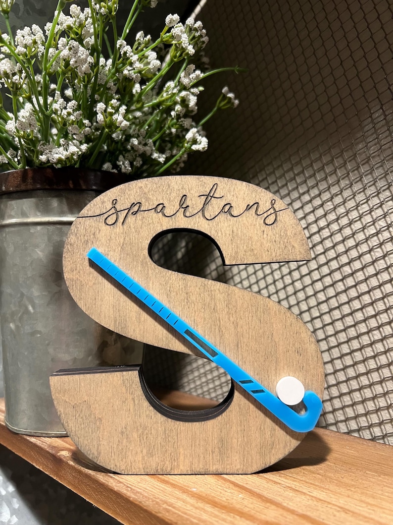 May include: Wooden letter S with the word "Spartans" carved into it. A blue hockey stick with a white puck is placed on the letter.