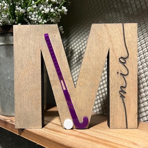 May include: Wooden letter M with a purple field hockey stick and a white ball. The letter is painted in a light brown wood stain. The letter has the name "mia" written in cursive on the right side.