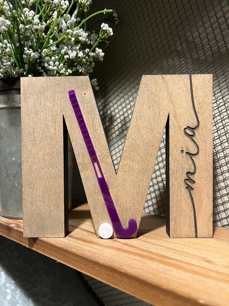 May include: Wooden letter M with a purple field hockey stick and a white ball. The letter is painted in a light brown wood stain. The letter has the name "mia" written in cursive on the right side.