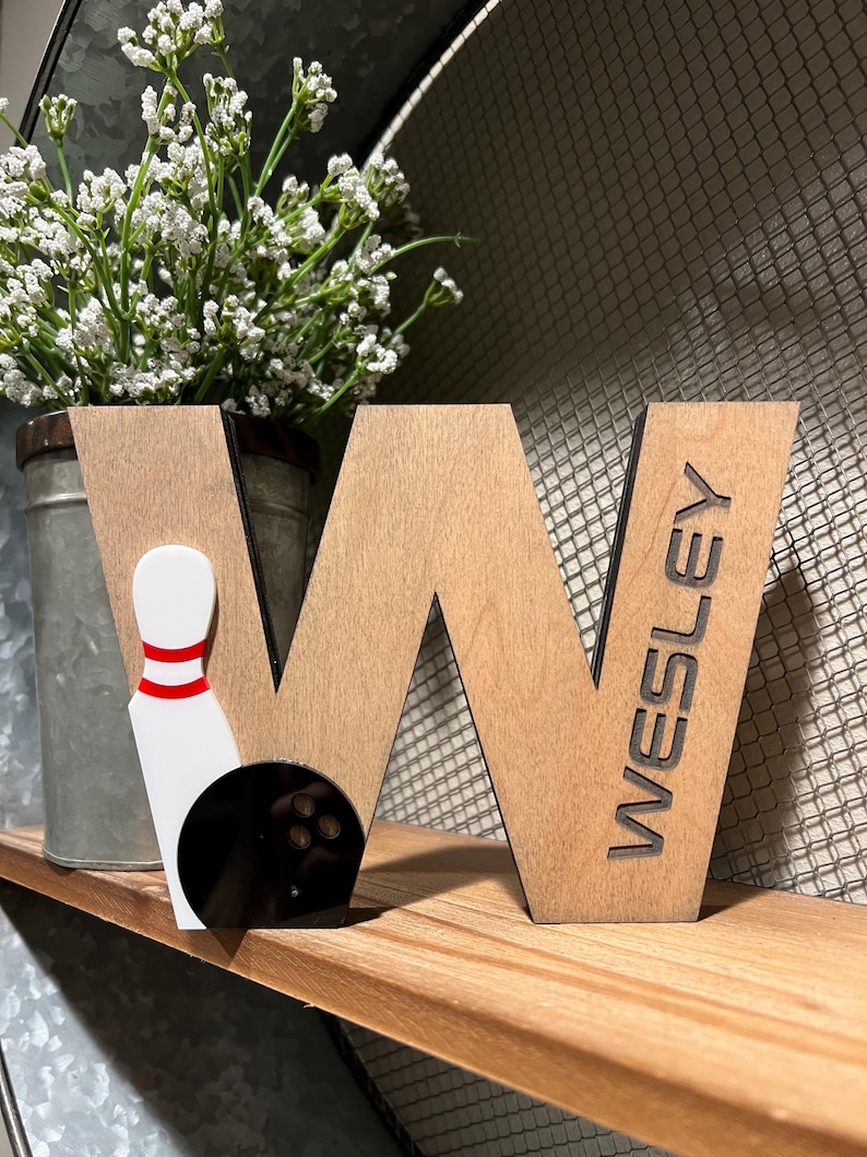 Pu&ograve; includere: Lettera W in legno con una palla e un birilli da bowling neri e bianchi attaccati. La lettera &egrave; macchiata di marrone e ha il nome "WESLEY" inciso sul lato destro.