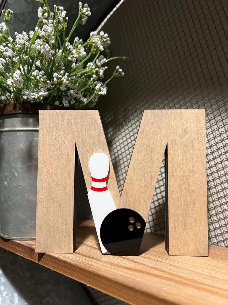 Pu&ograve; includere: Lettera M in legno con una palla da bowling nera e un biril bianco con strisce rosse.