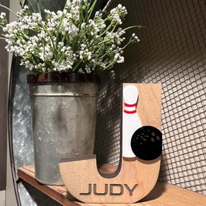 Pu&ograve; includere: Lettera J in legno con un design a forma di birilli e palla da bowling. La lettera &egrave; dipinta di marrone e ha il nome "JUDY" inciso nella parte inferiore.