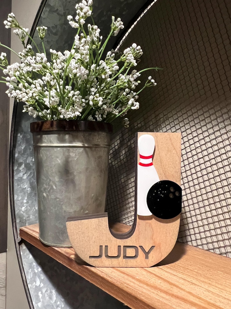 Pu&ograve; includere: Lettera J in legno con un design a forma di birilli e palla da bowling. La lettera &egrave; dipinta di marrone e ha il nome "JUDY" inciso nella parte inferiore.