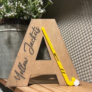 May include: Wooden letter A with the words "Yellow Jackets" engraved on it. A yellow field hockey stick is placed on the letter A.
